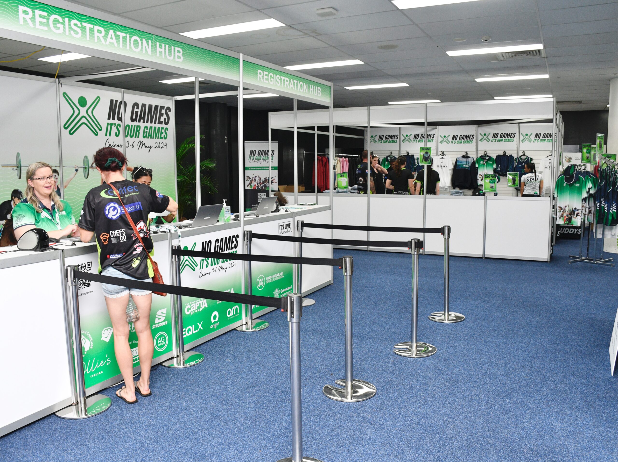 Registration hub at a convention with staff behind a white desk and attendees, green branding and banners reading 'NO GAMES IT'S OUR GAMES' along the counter edge.